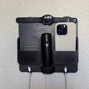 May include: Black wall-mounted organiser with compartments for a smartphone, smartwatch, and charging accessories. The organiser has a textured surface and includes a charging cable for the phone. The smartwatch is black with a black strap.
