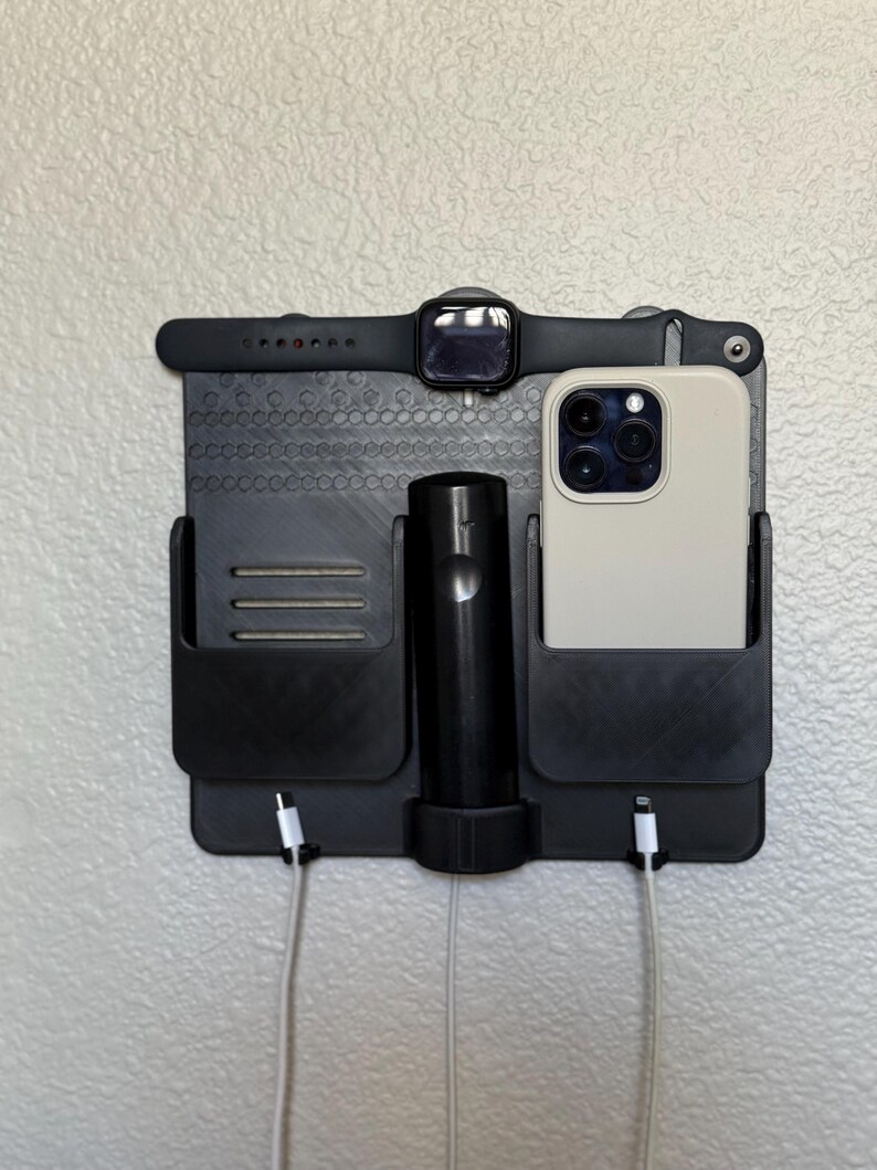 May include: Black wall-mounted organiser with compartments for a smartphone, smartwatch, and charging accessories. The organiser has a textured surface and includes a charging cable for the phone. The smartwatch is black with a black strap.