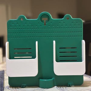 May include: A green and white wall-mounted organiser with two white pockets and a small bottom compartment. The organiser has a textured surface with a T-shaped hook at the top for hanging. The pockets have horizontal slots.