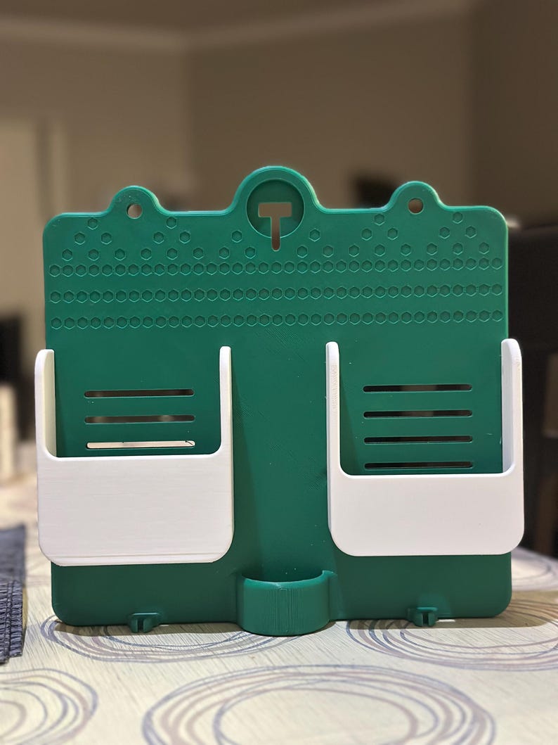 May include: A green and white wall-mounted organiser with two white pockets and a small bottom compartment. The organiser has a textured surface with a T-shaped hook at the top for hanging. The pockets have horizontal slots.