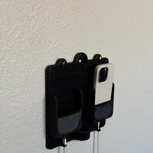 May include: A black wall-mounted phone and accessory organiser. It features two compartments, one holding a phone with a white case, and the other a small pocket. The organiser has a textured design and includes charging cables. Red and yellow accents are visible.