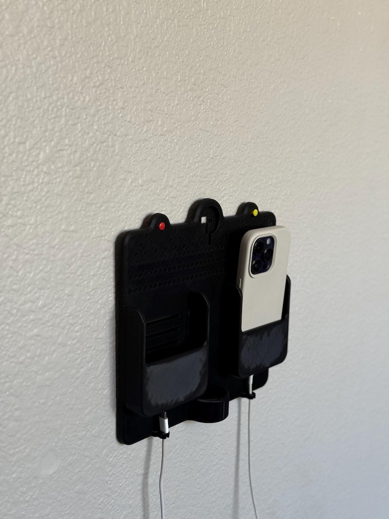 May include: A black wall-mounted phone and accessory organiser. It features two compartments, one holding a phone with a white case, and the other a small pocket. The organiser has a textured design and includes charging cables. Red and yellow accents are visible.