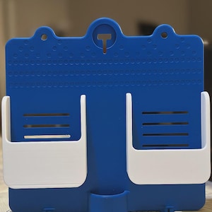 May include: A blue and white wall-mounted organiser with two white storage compartments and a small lower pocket. The blue surface has a textured pattern and a T-shaped cutout at the top.