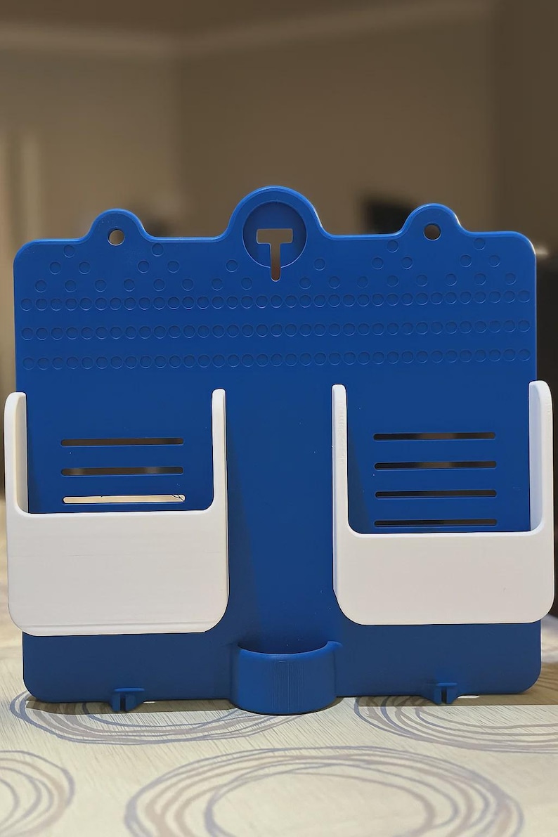 May include: A blue and white wall-mounted organiser with two white storage compartments and a small lower pocket. The blue surface has a textured pattern and a T-shaped cutout at the top.
