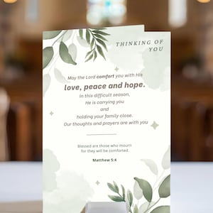 May include: A sympathy card with the text "Thinking of You" and a Bible verse. The card features watercolor-style green leaves and the words "love, peace and hope." The card is displayed on a white surface.