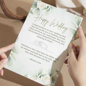 May include: A rectangular wedding card featuring white roses and green eucalyptus leaves. The text reads: Happy Wedding, Congratulations on your wedding day! May God guide you in building a home filled with love, joy, and peace. Genesis 2:24.
