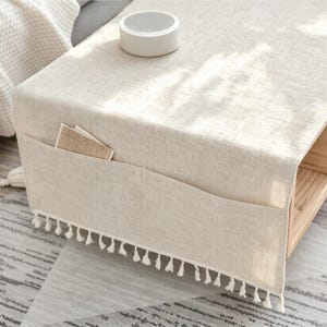 Op de afbeelding: Een beige tafelloper met linnenstructuur, voorzien van een zijvak en een decoratieve franje langs de onderrand. De loper ligt over een houten oppervlak en bevat een klein rechthoekig kaartje dat in het zijvak is gestoken.