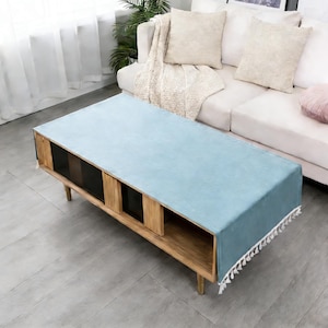 Peut inclure: Une table basse rectangulaire en bois avec un chemin de table bleu clair. La table a quatre pieds en bois et trois compartiments de rangement avec des portes en verre foncé. Le chemin de table a des glands blancs sur le bord.