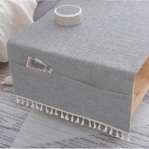 Könnte beinhalten: Eine graue Stoff-Couchtischabdeckung mit einer Tasche und weißem Quastenbesatz. Auf der Abdeckung liegt ein kleines, rundes, weißes Objekt. In der Tasche befindet sich eine Zeitschrift. Der Tisch ist aus Holz.