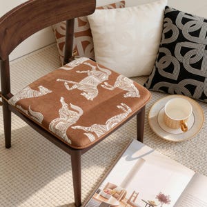 Puede incluir: Una silla de madera con un cojín de asiento marrón con un patrón de caballos blancos. Cojines decorativos con diseños geométricos y una taza de té sobre un platillo en el fondo. La silla está sobre una alfombra clara.