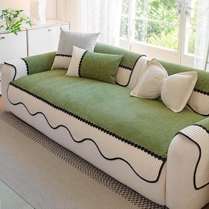 Puede incluir: Una funda de sofá verde con ribete negro ondulado y detalles de puntos negros. El sofá tiene una base blanca y está adornado con varios cojines decorativos en tonos verdes y blancos. La habitación tiene un ambiente luminoso y aireado con luz natural.