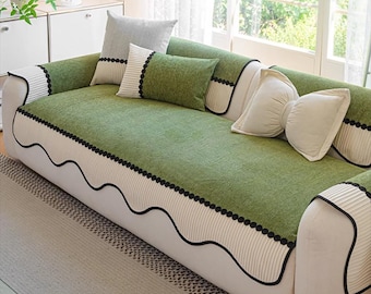 May include: A green sofa cover with a wavy black trim and black dot accents. The sofa has a white base and is adorned with several decorative pillows in shades of green and white. The room has a bright, airy feel with natural light.