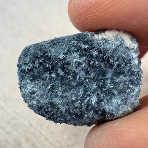 May include: A close-up of a dark blue mineral specimen with a crystalline structure. The mineral has a rough, textured surface with small, needle-like crystals. The edges of the mineral are a lighter color, with a white area on the top edge.