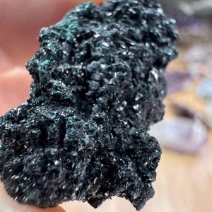 May include: A raw, dark mineral specimen with a highly textured, crystalline surface. The stone features deep black and charcoal grey tones with subtle, shimmering dark green highlights visible across its jagged, uneven, and reflective structure.