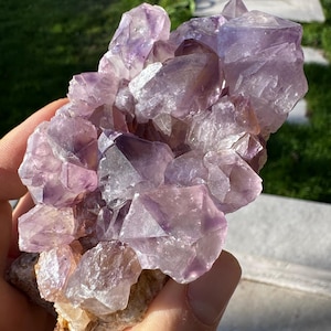 Könnte beinhalten: Nahaufnahme eines Amethyst-Kristallclusters, der verschiedene Lilatöne aufweist. Die Kristalle haben scharfe, geometrische Formen und sind auf einer rauen, beigefarbenen Matrix angeordnet. Das natürliche Licht betont die Facetten der Edelsteine.