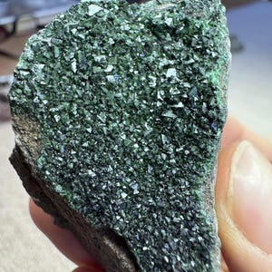 May include: A rough-hewn rock fragment with a dark brown base and a surface covered in sparkling, dark green crystals. The crystals have a metallic sheen, catching the light and creating a textured appearance. The mineral specimen is held in a hand.