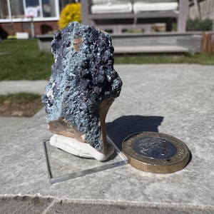 May include: A rough, textured mineral specimen with shades of blue, green, and purple. The rock is displayed on a clear acrylic stand, next to a coin. The coin is approximately 1 inch in diameter. The background is a sunny outdoor scene.