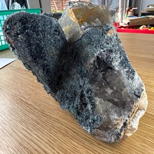 May include: A large, raw mineral specimen with a rough, textured surface. The stone features a mix of colours, including dark teal, grey, and translucent tan. The mineral is displayed on a wooden surface.