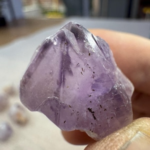 Könnte beinhalten: Ein roher Amethystkristall mit einer unregelmäßigen, facettierten Form. Der Kristall zeigt eine Abstufung von lilafarbenen Farbtönen, von hellem Lavendel bis zu tiefem Violett, mit einigen dunklen Einschlüssen. Die raue Textur und die natürliche Form des Steins sind sichtbar.