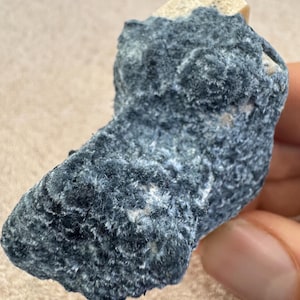 May include: A close-up of a mineral specimen featuring a dark blue, textured surface with a crystalline structure. The mineral has a rough, uneven shape and a light beige section at the top. The specimen is held in a hand.