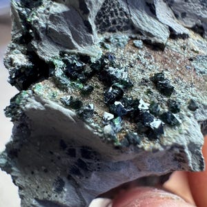 May include: Close-up of a mineral specimen showing a rough, grey rock formation. The rock is speckled with clusters of small, dark green and black crystals. The surface has a textured appearance, with areas of brown and grey.