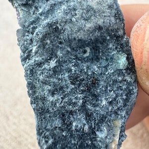 May include: A close-up of a rough, dark blue-green mineral specimen. The surface has a textured, fibrous appearance with small, crystalline formations. The mineral's color varies from deep blue to lighter green hues.