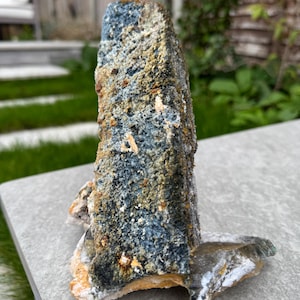May include: A natural, textured mineral specimen with a blue-grey core and a rough, multi-toned surface. The mineral has a unique, irregular shape and is displayed on a grey stone surface. The colors include shades of blue, grey, brown, and hints of orange.