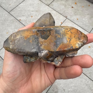 May include: A large, raw quartz crystal cluster with a smoky gray hue and areas of golden-brown oxidation. The crystal is held in a hand, showcasing its natural, unpolished form. The stone has a rough, textured surface.