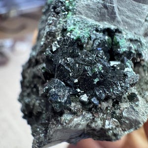 May include: A close-up of a rough, gray rock formation with dark, crystalline structures. The crystals are a deep, reflective black with hints of green and blue, creating a textured, mineral appearance. The rock's surface is uneven and detailed.