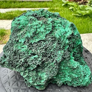 Könnte beinhalten: Ein großes Malachit-Exemplar mit rauer Textur. Der Stein weist eine lebendige grüne Farbe mit dunkleren, fast schwarzen Bereichen auf. Die Oberfläche des Minerals hat ein geschichtetes, faseriges Aussehen, das seine natürliche Entstehung hervorhebt. Der Malachit wird auf einer dunklen Oberfläche präsentiert.