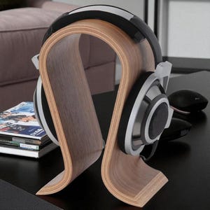 Puede incluir: Un soporte de madera para auriculares con un diseño curvo, que sostiene un par de auriculares negros, plateados y blancos. El soporte es de madera marrón claro y se encuentra sobre una superficie negra. También se ven una pila de revistas y un ratón de ordenador negro.