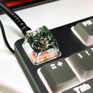 Może przedstawiać: Zbliżenie niestandardowej nasadki klawisza klawiatury. Przezroczysta nasadka klawisza zawiera zielony i złoty brokat oraz inne małe elementy dekoracyjne. Nasadka klawisza znajduje się na czarnej klawiaturze z szarymi klawiszami.