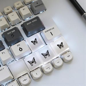 Może przedstawiać: Zbliżenie klawiatury komputera z niestandardowymi nakładkami na klawisze. Nakładki są białe i szare, z czarnymi motywami motyli i różnymi symbolami. Niektóre klawisze mają tekst, np. "QOJT BAIR" i "BHEEM SVIRLE".