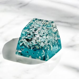 Może przedstawiać: Turkusowy, prostokątny klawisz klawiatury z żywicy z wtrąceniami srebrnej folii. Klawisz ma błyszczące wykończenie i jest przezroczysty. Leży na białej marmurowej powierzchni, tworząc kontrast kolorów.