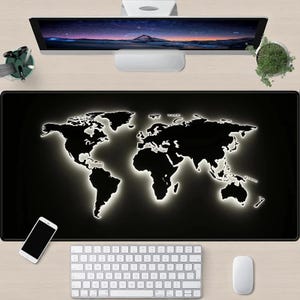 May include: A black desk mat featuring a world map with a glowing white outline. A white keyboard, mouse, and smartphone are visible. A computer monitor and a small plant are also on the desk.