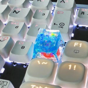 May include: Close-up of a mechanical keyboard with white keycaps. A custom keycap with a blue, red, and yellow resin design is in the center. The keyboard has a backlight, and the keycaps are labeled with numbers and symbols.