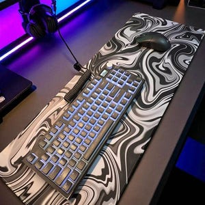 May include: A black and gray mechanical keyboard with blue backlit keys rests on a matching mousepad. The mousepad features a swirling, abstract design in shades of gray and white. A black computer mouse sits on the mousepad.