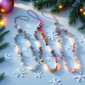 Può includere: Tre ciondoli per telefono con perline con diverse combinazioni di colori. Uno è grigio e blu, un altro multicolore e il terzo viola e rosa. Ogni ciondolo ha un anello per attaccarlo a un telefono. I ciondoli sono su una superficie azzurra con decorazioni a fiocco di neve.