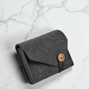 Puede incluir: Una cartera de fieltro gris oscuro con cierre de solapa y un botón de madera. La cartera se cierra con una cuerda marrón enrollada alrededor del botón. El fondo es una superficie de mármol blanco.