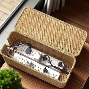 Puede incluir: Un organizador de cables rectangular de mimbre tejido con tapa con bisagras. En el interior, se ven dos regletas blancas con múltiples enchufes, junto con cables y enchufes eléctricos negros y blancos. El organizador está sobre una superficie de madera.