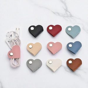 May include: A collection of heart-shaped cable organizers in various colors, including black, red, and pink. Each organizer has a snap closure and is shown with white earbuds. The organizers are displayed on a white marble surface.
