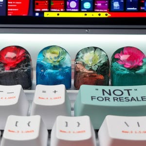 Może przedstawiać: Cztery niestandardowe, rzemieślnicze nakładki na klawisze z kwiatowymi wzorami w żywicy, z czerwonymi, niebieskimi i różowymi kwiatami. Nakładki na klawisze znajdują się na białej klawiaturze z jasnoniebieskim klawiszem z napisem "NOT FOR RESALE".