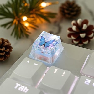 Może przedstawiać: Przezroczysty, kwadratowy klawisz klawiatury z niebieskim motylem. Klawisz znajduje się na białej klawiaturze, z widocznym klawiszem "Tab". W tle dekoracyjne szyszki i podświetlana girlanda.