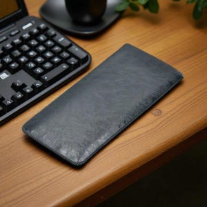 Può includere: Una custodia in pelle grigio scuro, probabilmente per un tablet o un laptop, è appoggiata su una scrivania di legno. La custodia ha un bordo cucito e una superficie strutturata. Una tastiera nera e una tazza nera su un piattino sono visibili.