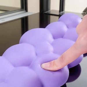 Può includere: Primo piano di un cuscino a forma di nuvola color lavanda. Il cuscino ha una superficie strutturata e a bolle. Un dito preme sul materiale morbido, mostrando la sua flessibilità. Il cuscino è progettato per il comfort e il supporto.