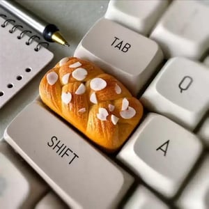 May include: A close-up of a keyboard with a loaf of bread on top. The bread is golden brown with white almond slices. The keyboard keys are white with black lettering, including "TAB", "SHIFT", "Q", and "A". A pen and notepad are in the background.