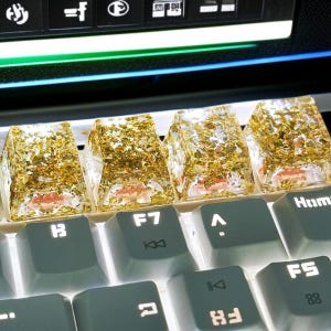 Może przedstawiać: Cztery przezroczyste żywiczne nakładki na klawisze klawiatury wypełnione płatkami złota, spoczywające na szarej klawiaturze. Nakładki na klawisze są prostokątne o trapezowym kształcie. Klawisze klawiatury poniżej są szare z białymi literami.