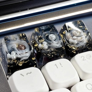 Może przedstawiać: Zbliżenie niestandardowych nakładek na klawisze klawiatury. Nakładki są przezroczyste z czarno-białym efektem marmuru, złotymi płatkami i osadzonymi perłami. Widoczne są również białe klawisze z symbolami "~", "1!" i "2@".