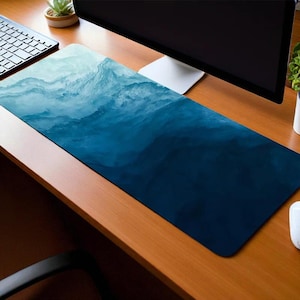 Puede incluir: Una alfombrilla de escritorio larga y rectangular con un diseño degradado. La parte superior presenta un patrón de olas azul claro y blanco que se transforma en un azul marino profundo en la parte inferior. La alfombrilla está sobre un escritorio de madera, con teclado, monitor y ratón.