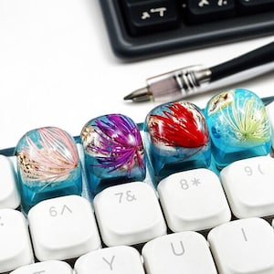 May include: Four keyboard keycaps with clear resin tops, each with a unique design. The keycaps feature pink coral, white shells, purple plant, and red plant. The keycaps are on a keyboard with white keys, with a calculator and pen in the background.